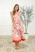 A woman standing in an indoor setting wearing a white and red orange floral tiered dress with a round neckline and short sleeves.