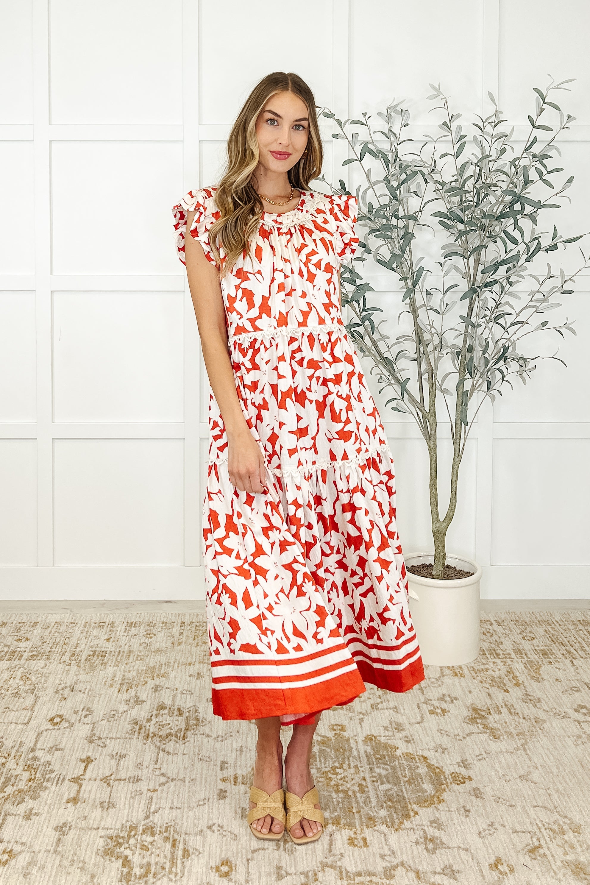 A woman standing in an indoor setting wearing a white and red orange floral tiered dress with a round neckline and short sleeves.
