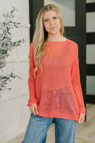 A woman wearing a coral loose knit sweater with a boat neckline and long sleeves.