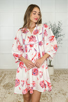 A woman standing in an indoor setting wearing a white floral dress with a v-neckline, puff sleeves, and side pockets.