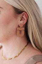 Brown earrings with a round top and geometric drop design, worn by a person.
