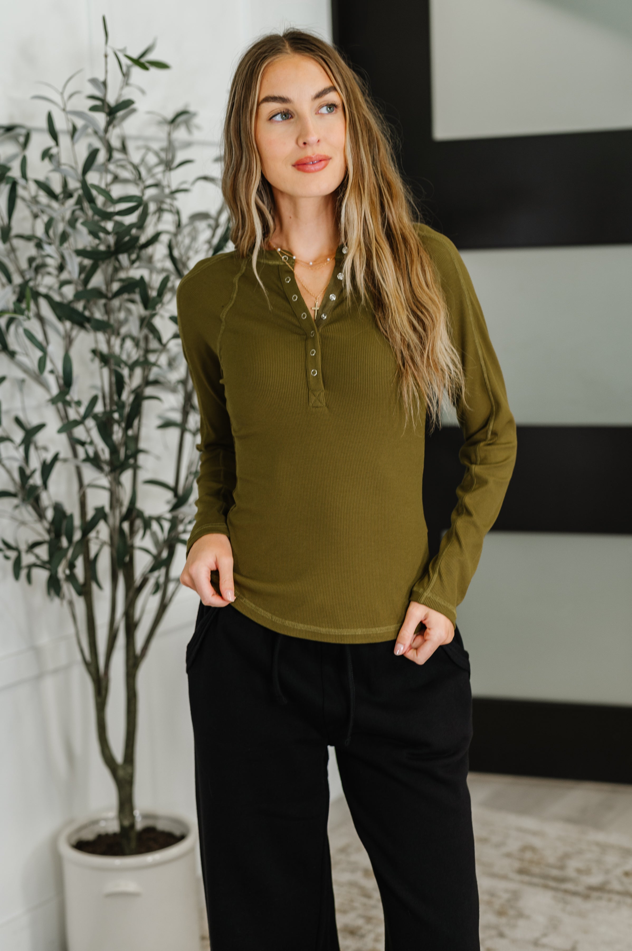 Woman wearing an olive green long-sleeve top and black pants indoors.