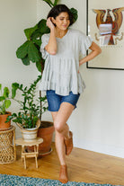 Woman in a gray ruffled top and blue shorts standing in a room with plants and a framed picture on the wall.