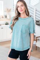 A woman wearing a light blue mineral wash top with a round neckline, dolman sleeves, and a raw hem patch pocket.
