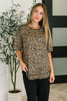 Woman wearing a leopard print top and black pants standing indoors with a plant in the background.