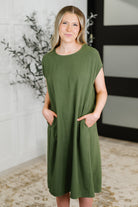 Woman wearing an olive green dress standing indoors with a decorative plant in the background.