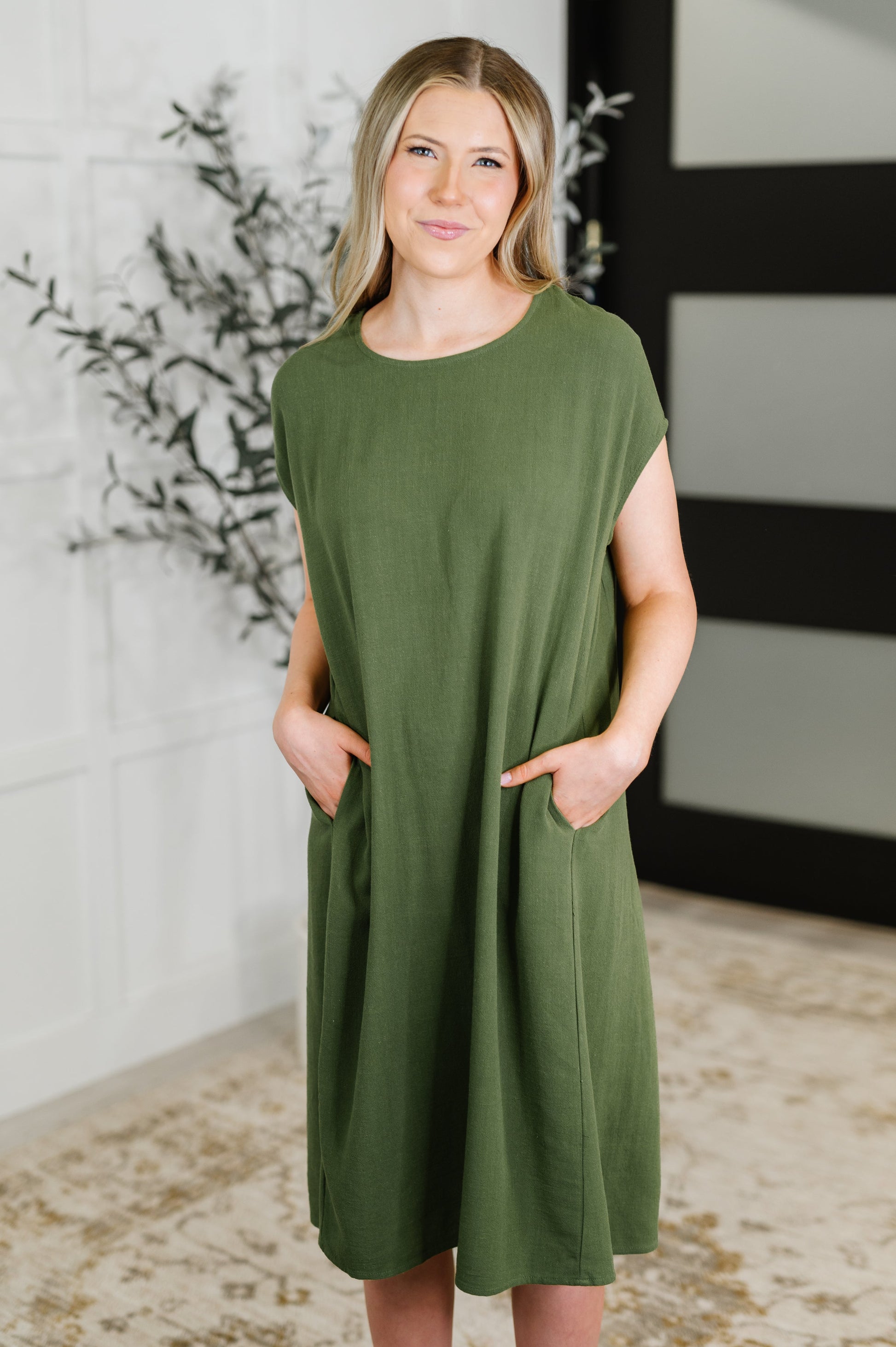 Woman wearing an olive green dress standing indoors with a decorative plant in the background.