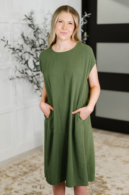 Woman wearing an olive green dress standing indoors with a decorative plant in the background.