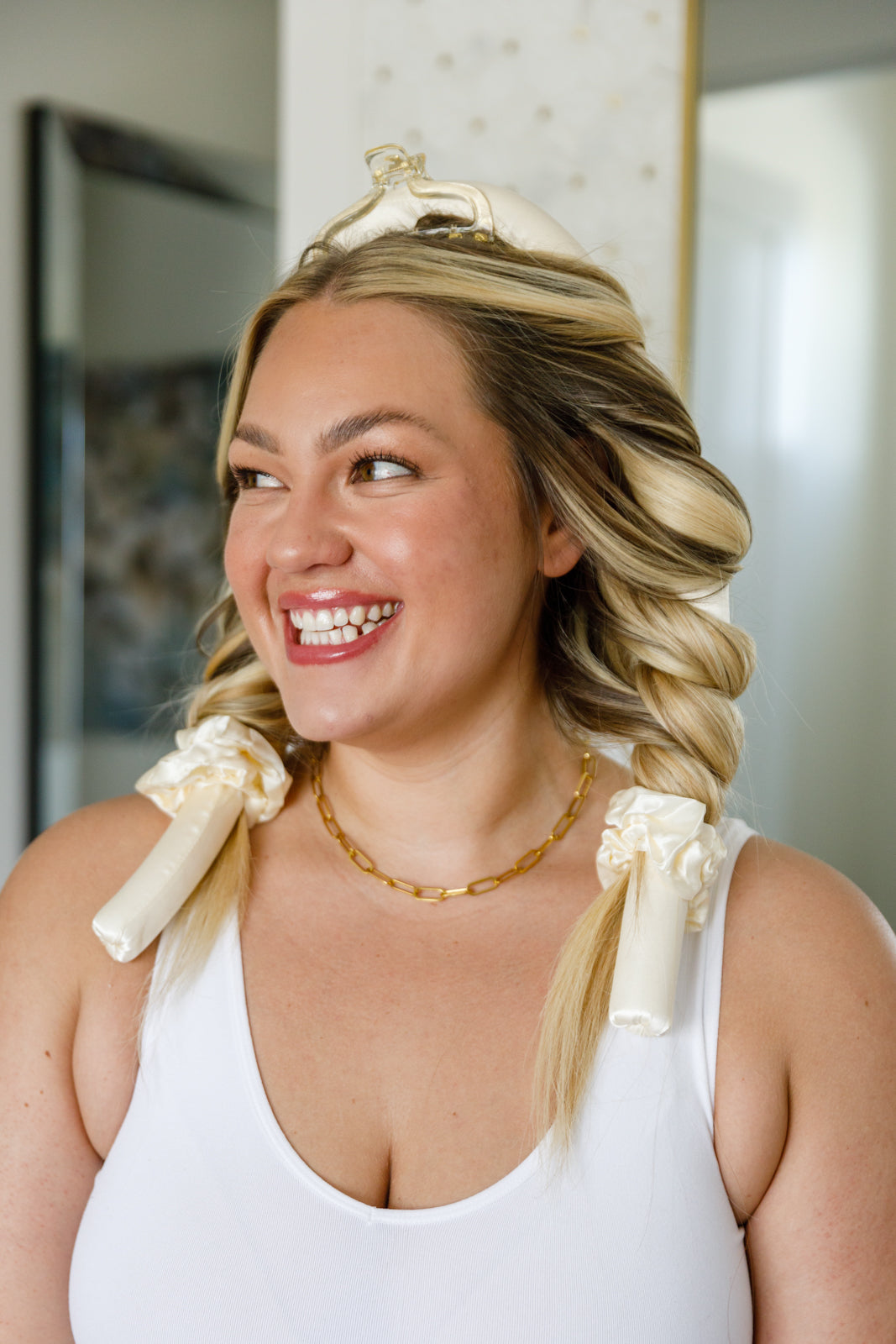 Woman using heatless hair curlers, wearing a white tank top indoors.