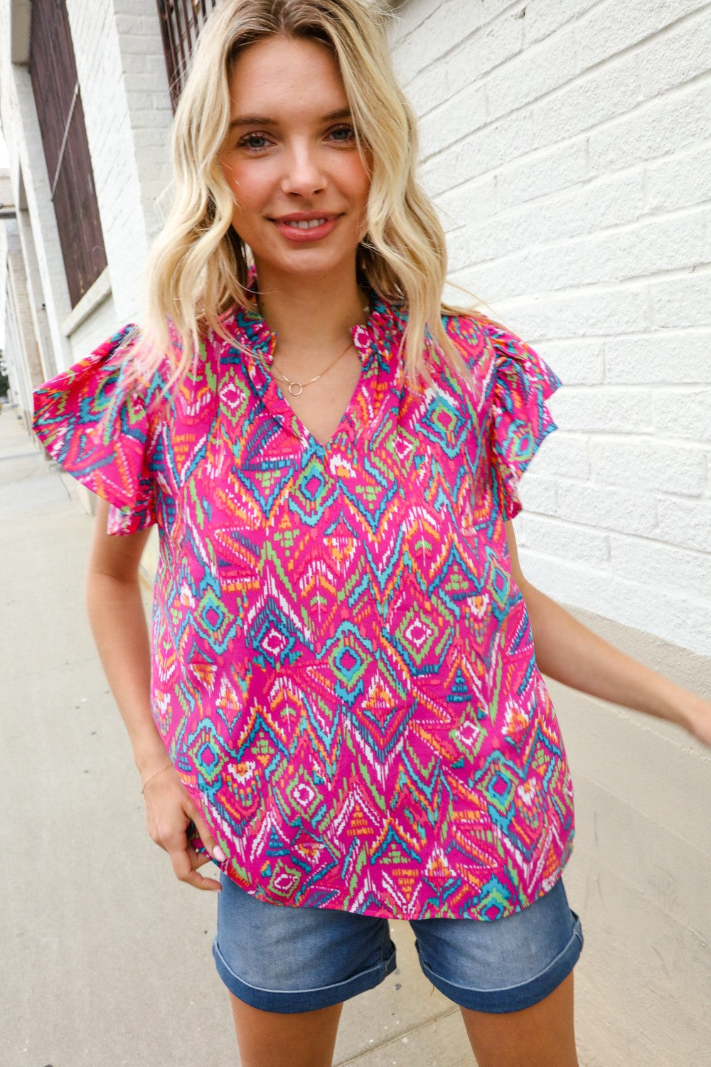 A woman wearing a fuchsia abstract print notch frill neck blouse with short puff sleeves, paired with denim shorts.