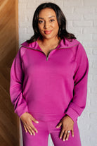 A woman standing in a room with a white brick wall, wearing a magenta half zip pullover with mock neck and raglan sleeves, paired with matching pants.