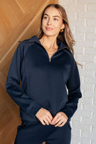 A woman standing in a doorway wearing a navy blue travel half zip pullover with raglan sleeves and a mock neckline.