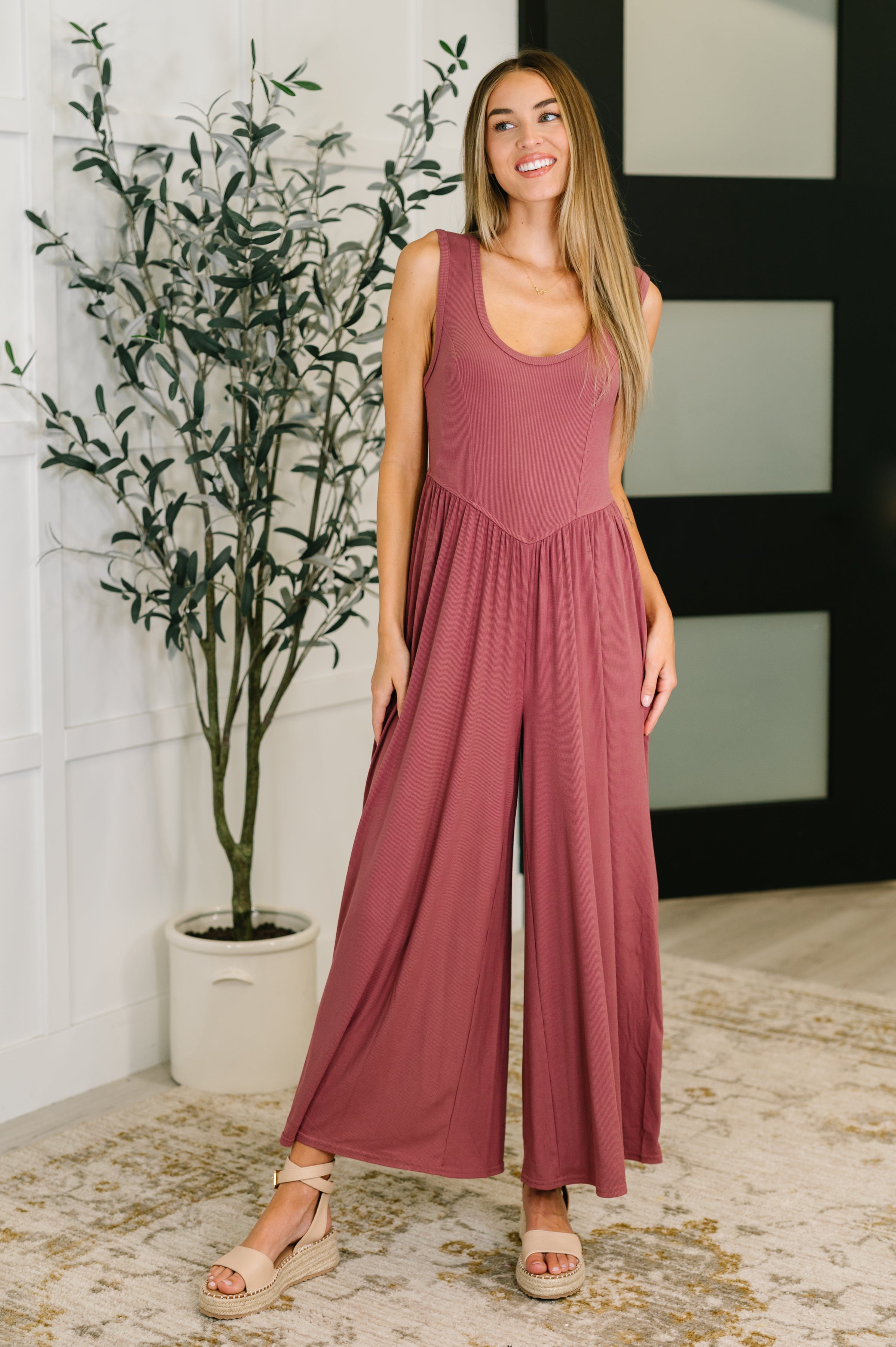 Woman wearing a mauve jumpsuit standing indoors with a plant in the background