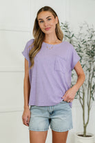 A woman wearing a lavender basic tee with a round neckline, short dolman sleeves, and a front pocket, paired with denim shorts.