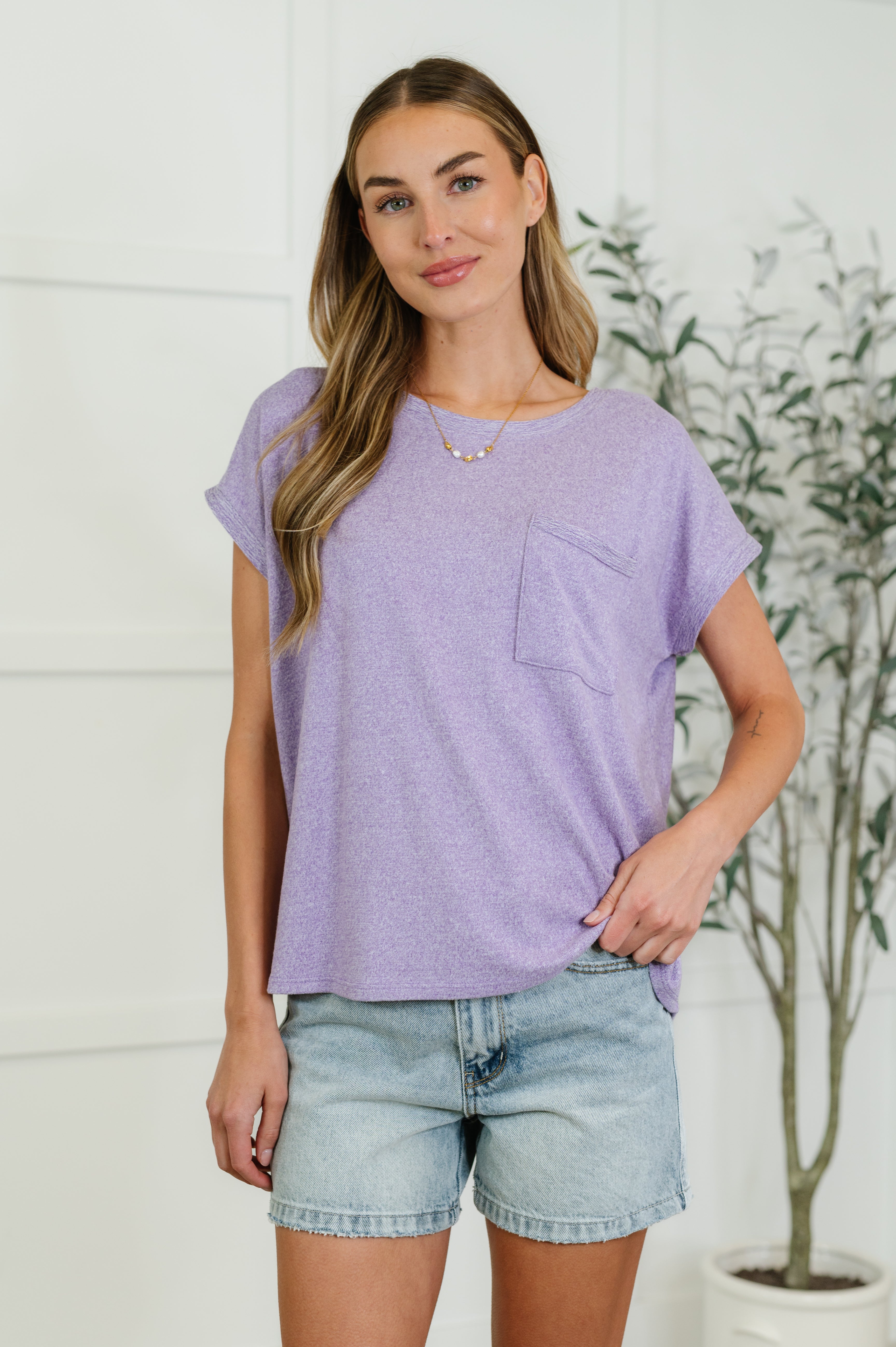 A woman wearing a lavender basic tee with a round neckline, short dolman sleeves, and a front pocket, paired with denim shorts.