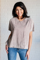 Woman wearing a beige t-shirt and blue jeans against a white brick wall.