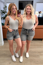 Two woman wearing high-rise button-fly cuffed shorts in grey, with one model holding a phone in front of the other. Both are wearing white sneakers and jewelry.