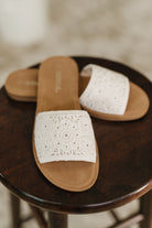 Women's white sandals with brown soles on a wooden stool.