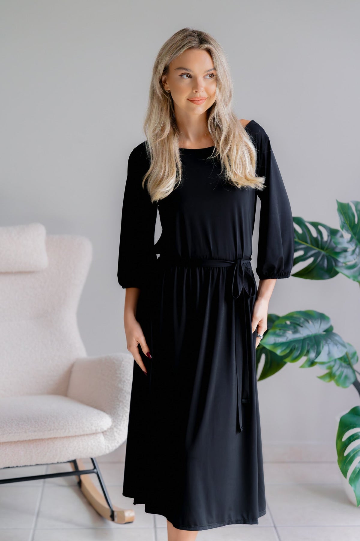 Woman wearing a black dress with a boat neckline and 3/4 sleeves, standing indoors.