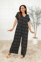 A woman standing in a room wearing a black and multicolor dotted jumpsuit with a V-neckline and wide legs.