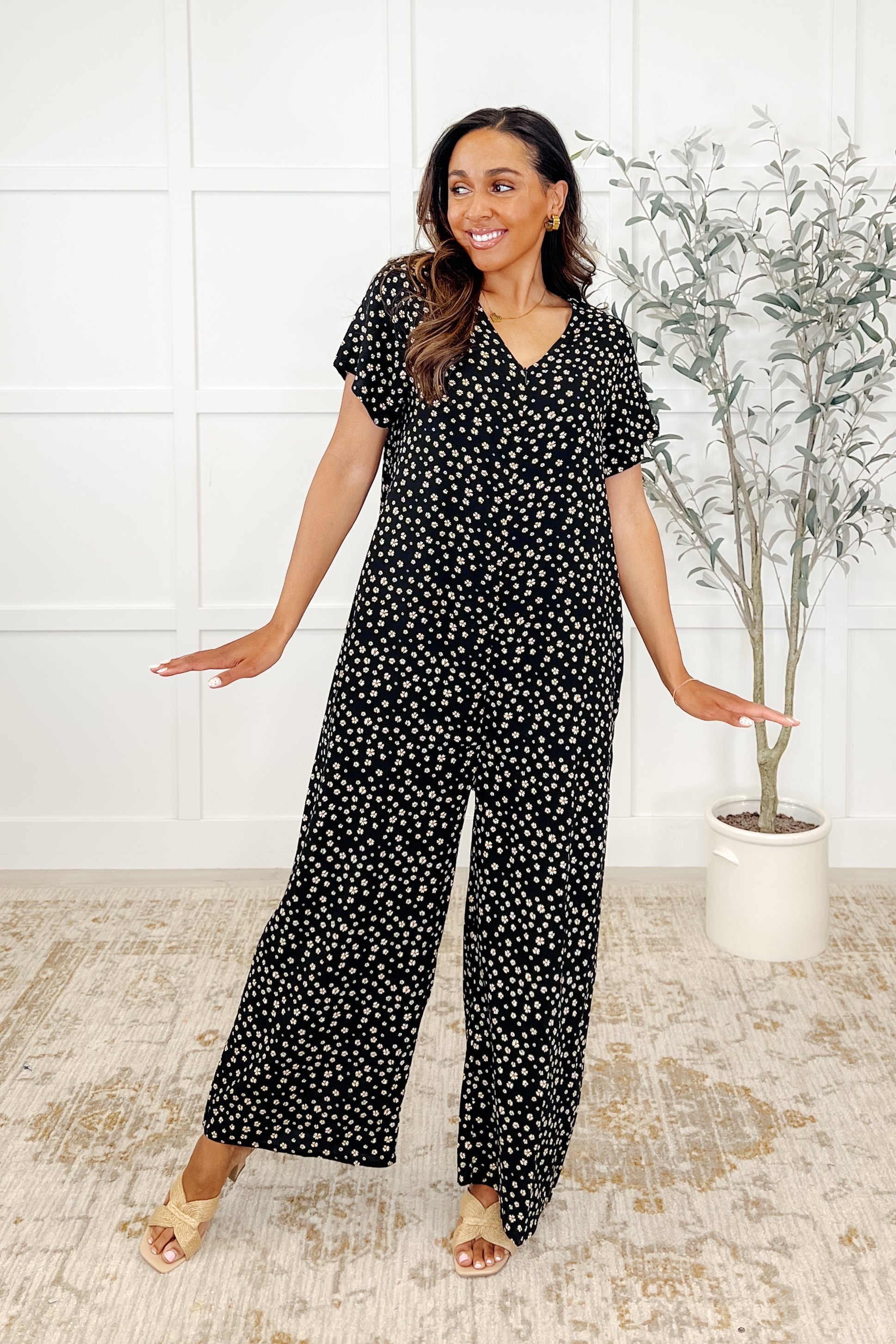 A woman standing in a room wearing a black and multicolor dotted jumpsuit with a V-neckline and wide legs.