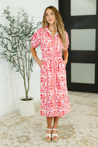 Woman wearing a red and white patterned dress standing indoors with a plant in the background.