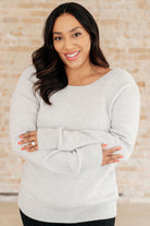 A woman wearing a light gray sweater with a boat neckline and ribbed cuffs.