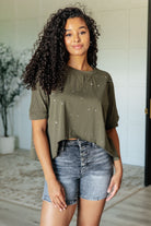 A woman wearing an olive green asymmetrical distressed top with a round neckline and paint splatter details, paired with denim shorts.