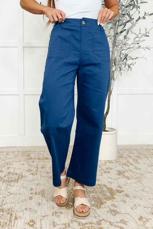 A woman standing against a white background wearing royal blue straight leg trousers with a button and zipper fly, functional front and back patch pockets, and a cropped wide leg.