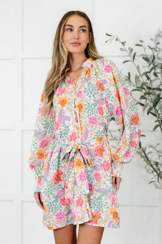 A woman wearing a floral dress with a round neckline, full button closure, gathered shoulders, and a waist tie.