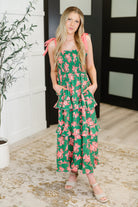 Woman wearing a green floral dress standing indoors with a plant and door in the background.