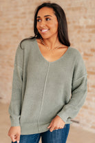 Woman wearing a sage green V-neck sweater with ribbed cuffs and hem.
