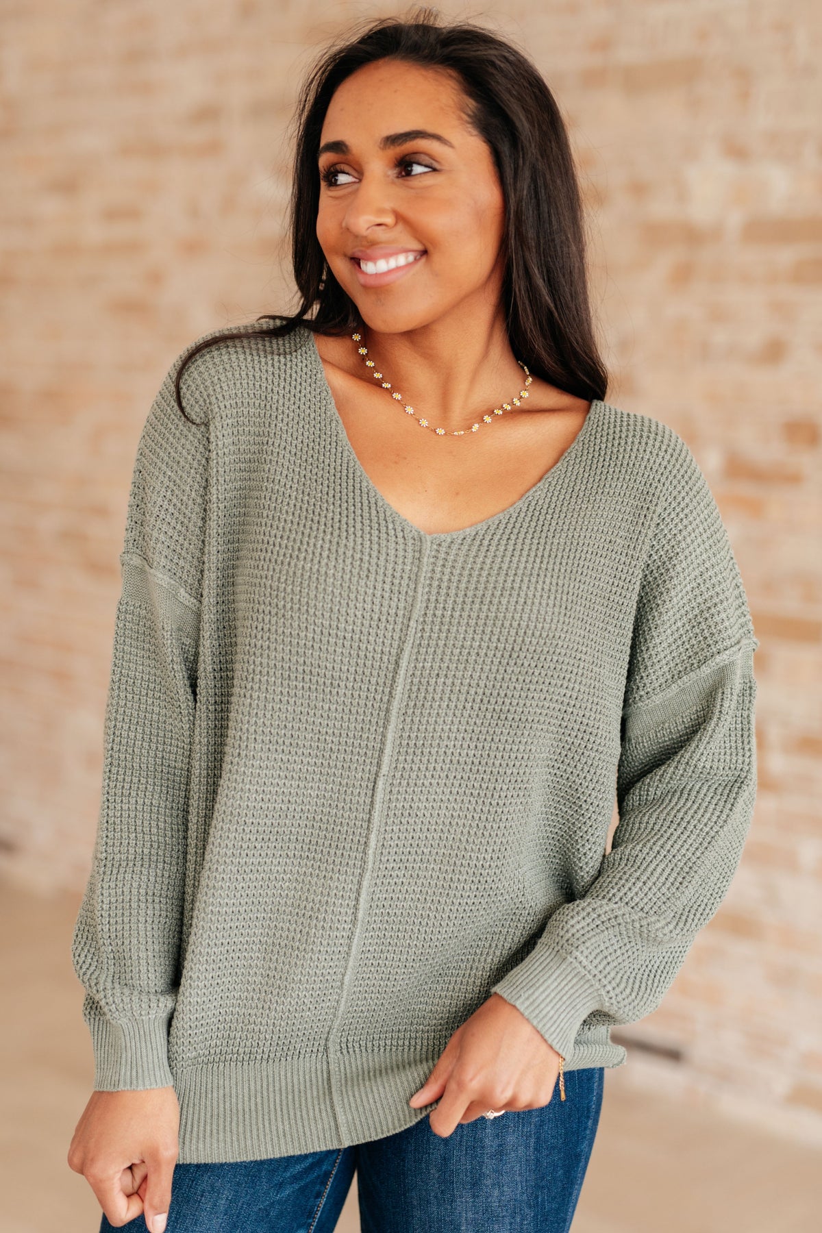 Woman wearing a sage green V-neck sweater with ribbed cuffs and hem.