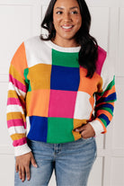A colorful checkered color block sweater with a ribbed round neckline and drop shoulders, modeled with jeans.