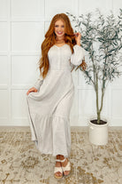 A woman standing in a room with a white dress featuring a v-neckline, balloon sleeves, and a tiered hem. She has long wavy hair and is wearing open-toe heeled sandals.