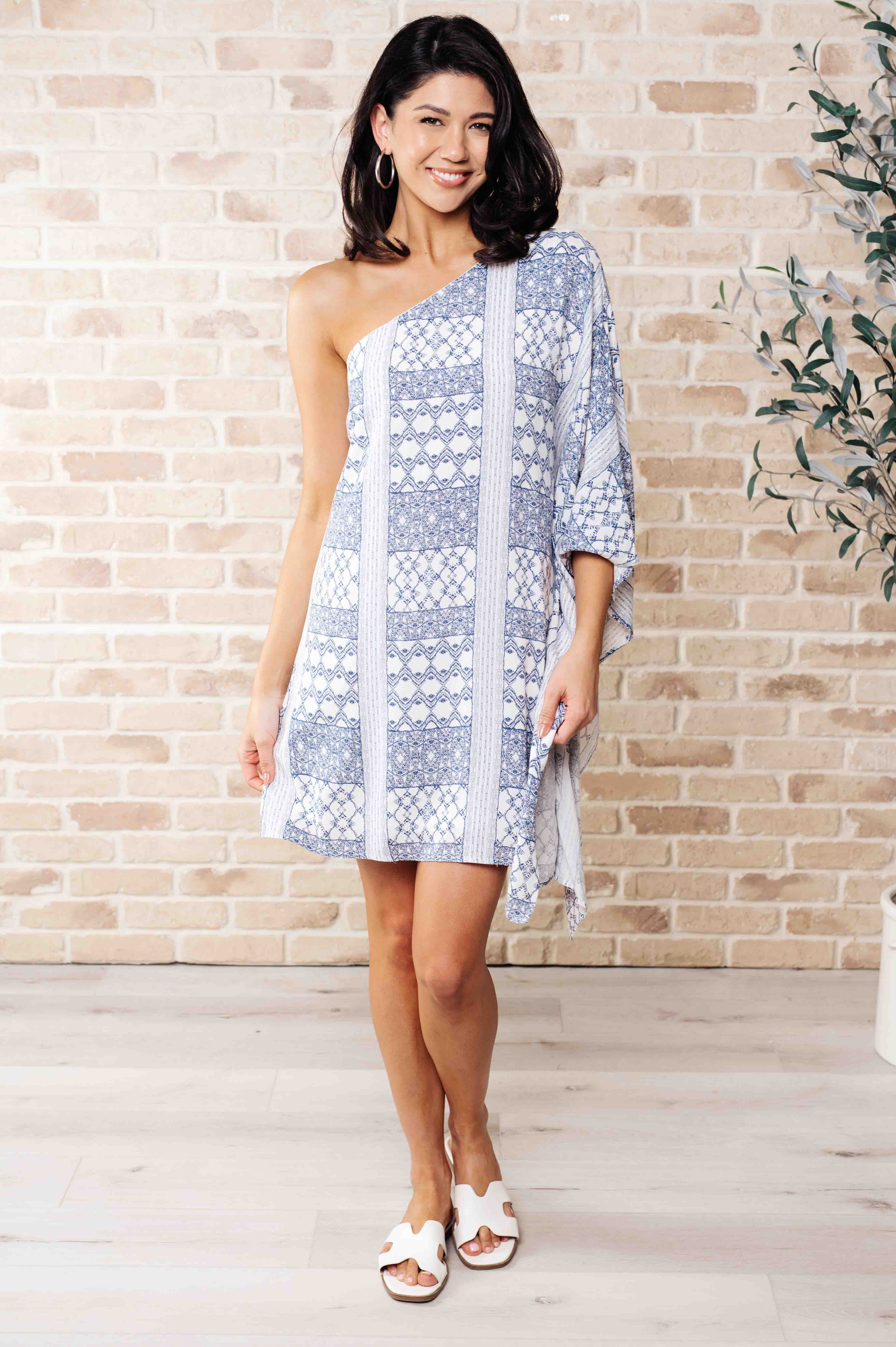 A woman wearing a blue and white one-shoulder mini dress with a geometric pattern, paired with white sandals.