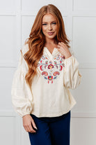 Woman wearing a cream blouse with floral embroidery against a white tiled wall.