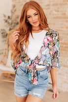 A woman wearing a multicolored floral front tie blouse with flutter sleeves and a scooped hem, paired with a white top and blue denim shorts.