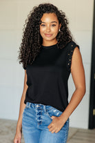 A woman wearing a black knit top with a round neckline and cap sleeves with stud embellishments, paired with blue jeans.