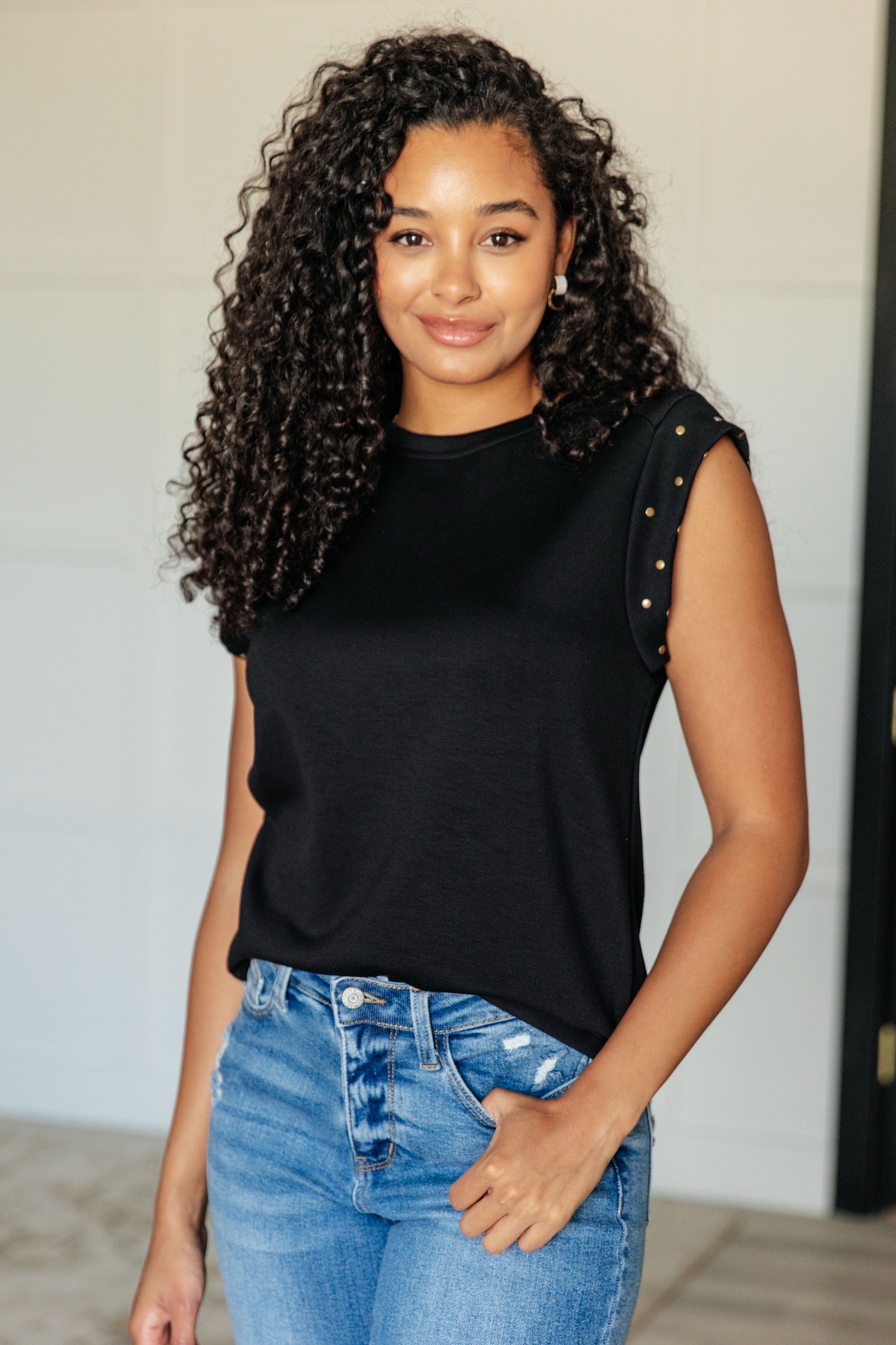A woman wearing a black knit top with a round neckline and cap sleeves with stud embellishments, paired with blue jeans.