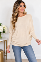A woman wearing a sand beige dolman sleeve boat neck top with a relaxed, boxy silhouette.