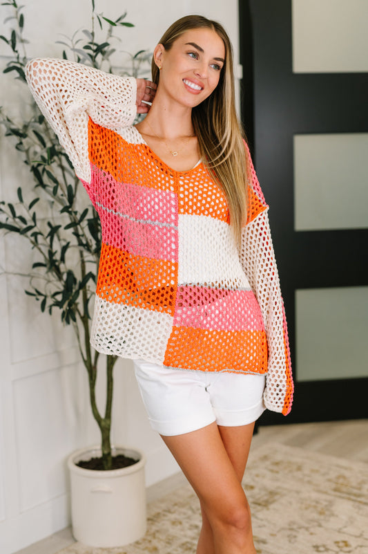 Woman wearing a colorful crochet top and white shorts standing indoors with a plant in the background.