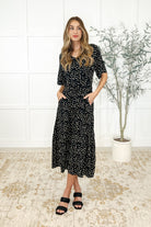 Woman wearing a black dress with a floral pattern, featuring a scooped neckline, three-quarter sleeves, side pockets, and an attached waist tie.