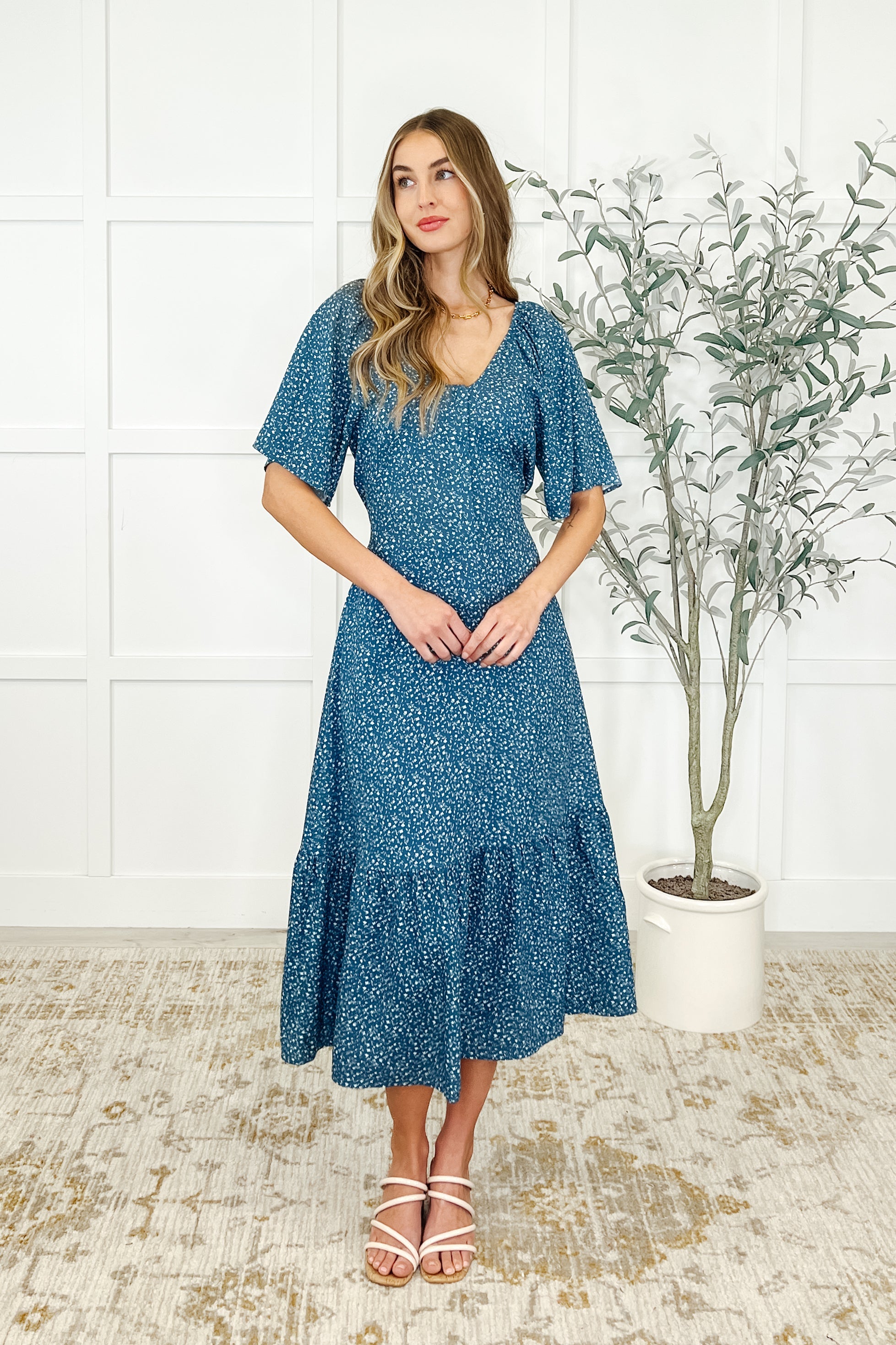 Woman wearing a blue floral dress with a scooped neckline, raglan sleeves, and tiered details, paired with strappy sandals.