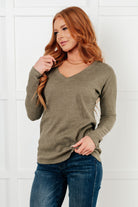 Woman wearing an olive green sweater and blue jeans against a white tiled wall.