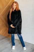 A person wearing an oversized black velour blanket hoodie with a hood and balloon sleeves, standing indoors.