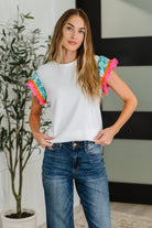 Woman wearing a white t-shirt with colorful sleeves and blue jeans indoors.