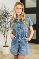 Woman wearing a denim romper standing indoors with a plant in the background