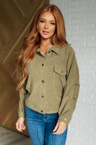 Woman wearing an olive green button-up shirt and blue jeans against a white brick wall.