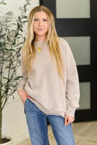 Woman wearing a beige sweatshirt and blue jeans standing indoors with a plant in the background
